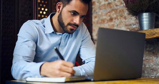 Man studying at home behind his laptop, and making notes on a pad